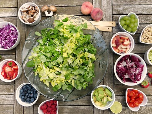 Hier sehen Sie eine große Schüssel mit grünen Salat. Um die Schüssel stehen viele kleine Schüsseln mit Zutaten, die man dem Salat hinzu geben kann. Zum Beispiel Tomaten, Nüsse, Zwiebeln, Beeren und noch vieles mehr. 
