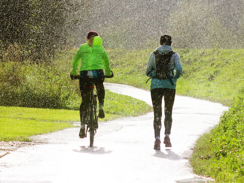 Hier sehen Sie zwei Personen draußen im Regen sport machen. Die eine Person fährt Rad, die ander Person joggt daneben. 