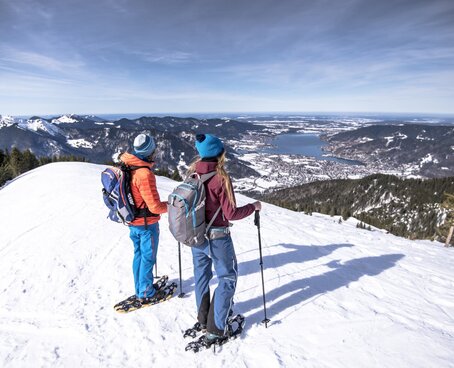 Hier sehen Sie zwei Personen beim Wandern auf Schneeschuhen. Sie haben den Gipfel erreicht und blicken auf die umliegenden Berge. 