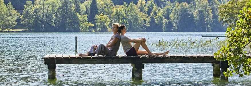Hier sehen Sie einen Steg von der Seite der in einen großen See läuft. Auf dem Steg sitzen eine Frau und ein Mann, Rücken an Rücken und genießen das schöne Wetter. Hinter ihnen sind Berge und Wald zu sehen. 