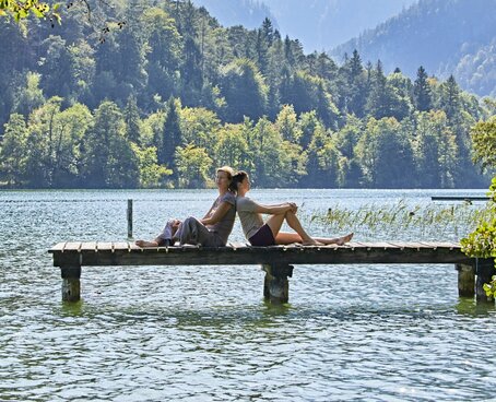 Hier sehen Sie einen Steg von der Seite der in einen großen See läuft. Auf dem Steg sitzen eine Frau und ein Mann, Rücken an Rücken und genießen das schöne Wetter. Hinter ihnen sind Berge und Wald zu sehen. 